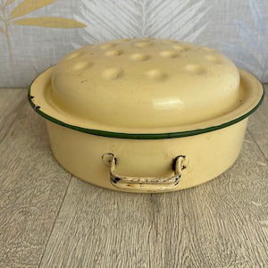 May include: A cream-coloured enamelware roasting pan with a lid. The lid features multiple small holes. The pan has a green trim and two handles. The pan is on a wooden surface.