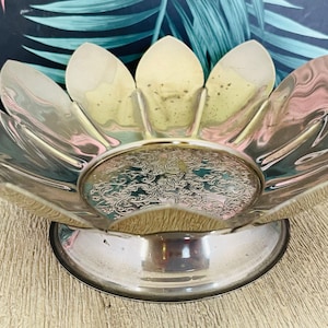 May include: A silver-coloured, flower-shaped serving bowl. The bowl has a pedestal base and a detailed, engraved design in the centre. The petals of the bowl are curved and pointed, creating a unique, decorative piece. The bowl is likely made of metal.