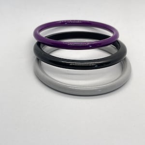 May include: Three stacked bangles in shades of purple, black, and grey. The bangles are smooth and glossy, with a simple, minimalist design. The bracelets are round and appear to be made of a solid material.