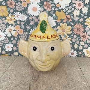 May include: A ceramic marmalade jar featuring a yellow, face-shaped base with two side handles and a matching lid. The lid is shaped like a lemon with a green leaf on top and the word MARMALADE printed in brown letters across the front surface.