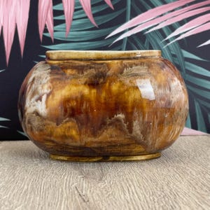 May include: A round, glazed ceramic pot with a wide opening. The pot features a blend of brown, tan, and white colors, creating a marbled effect. The base and rim are a lighter tan color. The pot sits on a wooden surface.