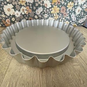 May include: A silver-colored tart pan with a fluted edge. The pan has a removable bottom and is designed for baking tarts and quiches. The pan is sitting on a wooden surface.