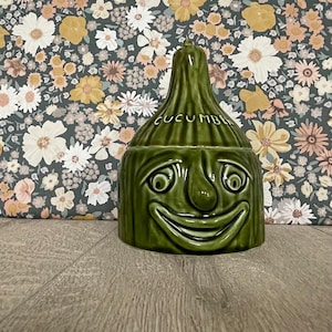 May include: A glossy dark green ceramic jar shaped like a stylised vegetable with a pointed lid. The jar features a moulded face with wide eyes and a broad smile, and the word CUCUMBER is embossed in white lettering on the upper section.