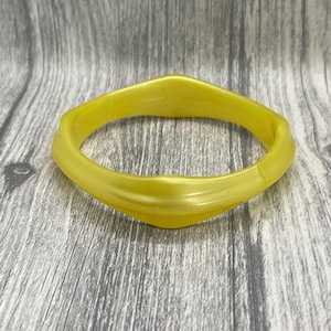 May include: A bright yellow bangle bracelet with a smooth, glossy finish and a distinct geometric, multi-faceted shape. The bracelet features a continuous, solid construction with slightly raised edges along the outer circumference of the band.