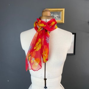 May include: A sheer red scarf with a floral pattern in yellow, orange, and blue is draped around a mannequin. The scarf is lightweight and appears to be made of silk or a similar fabric. The background is a dark gray wall with framed artwork.