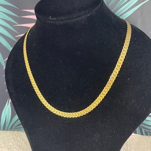 May include: A gold-colored, flat chain necklace displayed on a black velvet bust. The necklace has a herringbone design, with each link lying flat against the next. The background features a colorful floral pattern.