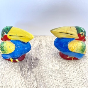 May include: A pair of toucan-shaped salt and pepper shakers. Each ceramic shaker features a yellow beak, green head, blue body, and red and yellow accents. The shakers are designed to resemble the tropical bird.
