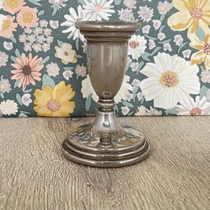 May include: A silver-colored candlestick holder with a round base and a flared top. The candlestick holder has a detailed design and is made of metal. The background features a floral pattern with yellow, white, and pink flowers.