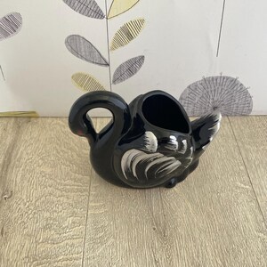 May include: A black ceramic swan-shaped planter with white brushstroke details on the wings and tail. The planter has a curved handle and a red accent on the beak. The interior is hollow, suitable for small plants or decorative items.