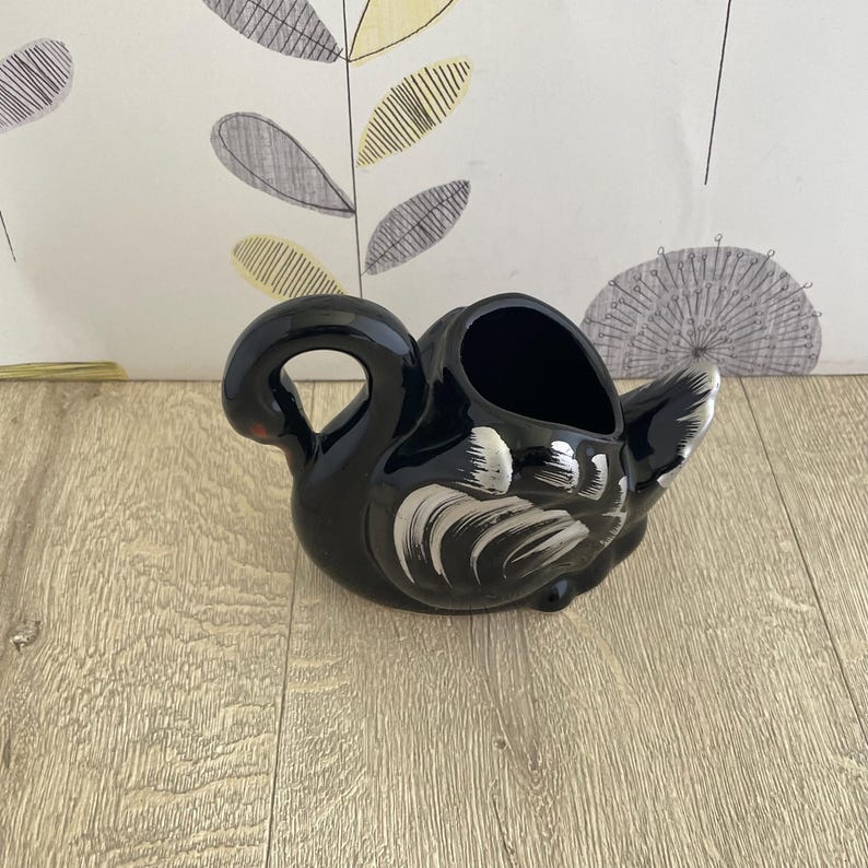 May include: A black ceramic swan-shaped planter with white brushstroke details on the wings and tail. The planter has a curved handle and a red accent on the beak. The interior is hollow, suitable for small plants or decorative items.