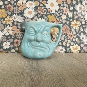 May include: A light blue ceramic creamer jug featuring a moulded face with a winking eye, a prominent nose, and a smirk. The jug has a curved handle on the right side and a small pouring spout at the top, showcasing a glossy finish.