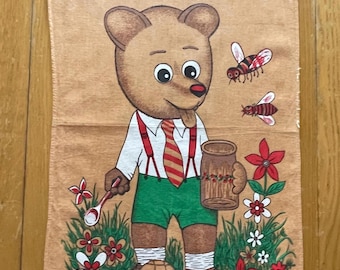 May include: Tan kitchen towel featuring a cartoon bear character wearing a white shirt, green shorts, and a red striped tie. The bear holds a spoon and a mug, surrounded by flowers and bees. A honey-comb is at the bear's feet.