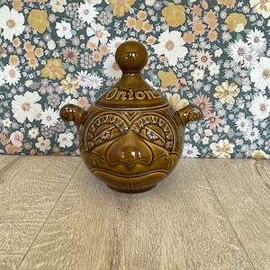 Könnte beinhalten: Eine braune Zwiebelsuppenterrine aus Keramik mit Deckel. Die Terrine hat ein gesichtsähnliches Design mit einer knolligen Nase und Augen. Das Wort "Onion" steht auf dem Deckel. Sie hat zwei Griffe und steht auf einer Holzoberfläche.