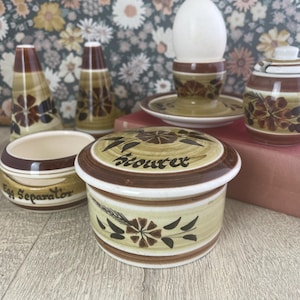 May include: A set of vintage ceramic kitchen items with a floral pattern in brown, beige, and white. The set includes salt and pepper pots, an egg separator, a lidded container with "Stoutex", a sugar bowl, and an egg cup with a plate.