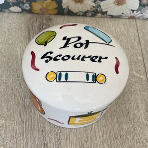 May include: A round white ceramic container with a lid featuring the hand-painted black text Pot Scourer. The surface is decorated with colourful illustrations of kitchen items including a green sponge, a blue brush, and yellow cookware.