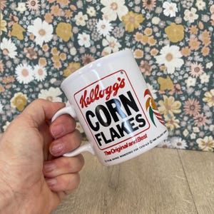 May include: White ceramic mug with the Kellogg's Corn Flakes logo in red and black. The mug has a curved handle and the text "The Original and Best". A colorful rooster graphic is also visible.