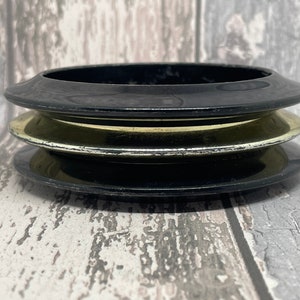 May include: Three stacked bangles, the top and bottom are black, the middle one is gold. The bangles have a smooth, glossy finish. They are set against a weathered, grey wooden surface. The bangles are likely for wearing as jewellery.