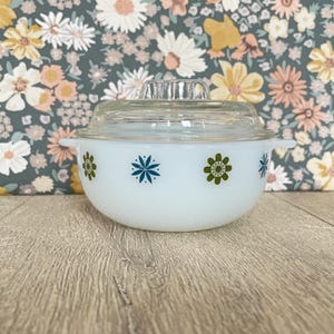 May include: A white Pyrex bowl with a clear glass lid. The bowl is decorated with blue and green floral designs. The bowl has two small handles on either side.