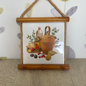 May include: A vintage kitchen wall hanging featuring a ceramic tile with a still life illustration of fruit, a jug, and a bottle. The tile is framed in wood and suspended by a rope. The bottom has three wooden pegs.