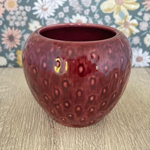 May include: A burgundy ceramic vase with a textured surface. The round vase has a wide opening and a glossy finish. The surface is covered in small, raised bumps, giving it a unique, tactile appearance. The vase is likely for decorative purposes.