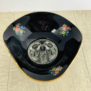 May include: A black, vintage flower arranging bowl with a gold rim and floral accents. The bowl has a clear glass flower frog in the center. The floral accents include red, yellow, blue, and pink flowers.