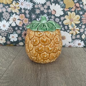 May include: A ceramic pineapple-shaped jar with a removable lid. The body of the jar features a textured, golden-brown pineapple pattern, while the lid is shaped like green pineapple leaves. The piece has a glossy finish and a rounded shape.