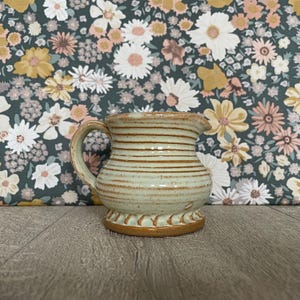 May include: A ceramic creamer pitcher with a light green glaze and horizontal brown stripes. The pitcher features a rounded body, a small pouring spout, a curved handle, and a decorative scalloped base with a natural brown clay finish.