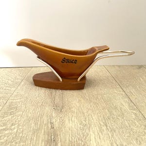 May include: A brown ceramic gravy boat with the word "Sauce" printed on the side. The boat is supported by a silver metal frame and a wooden base. The handle is also made of silver metal.