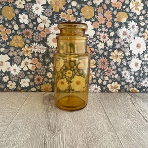 May include: A cylindrical amber glass storage jar with a matching glass lid. The jar features a smooth, translucent finish and a wide mouth opening. The glass is uniform in colour, showing a warm, golden-brown hue throughout the entire vessel.