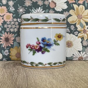 May include: A white porcelain jar with a lid, featuring a central floral arrangement of blue, yellow, and pink flowers. The jar is decorated with gold trim and a green leaf vine pattern along the top and bottom edges of the cylindrical body.