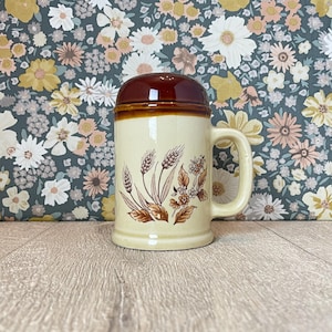 May include: A vintage beer stein with a brown lid and handle. The beige ceramic mug features wheat and floral designs in shades of brown. The stein is approximately 6 inches tall.