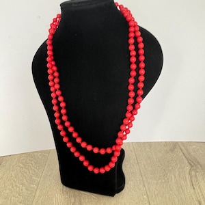 May include: A double-strand red beaded necklace displayed on a black jewellery bust. The beads are small and round, creating a continuous, vibrant line of colour. The necklace is draped elegantly, showcasing its length and design.