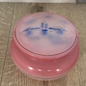 May include: A pink ceramic jar with a lid. The lid has a blue and white design of a sailboat on a body of water.