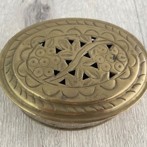 May include: Oval brass box with a decorative lid. The lid features a floral design with cut-out shapes. The box has a slightly aged appearance, suggesting it is vintage or antique. The box is sitting on a light-colored wooden surface.