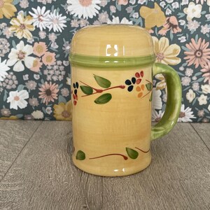 May include: A yellow ceramic beer stein with a green handle and rim. The stein is decorated with a floral design of green leaves and red, blue, and orange berries. The stein has a lid.