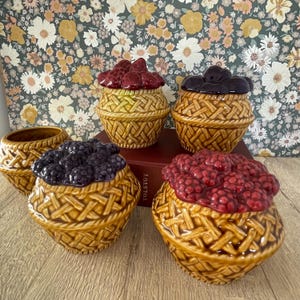 May include: A selection of ceramic berry jars with a woven basket design. The lids are shaped and coloured to resemble strawberries, blackberries, and raspberries. The jars are brown and sit on a wooden surface.