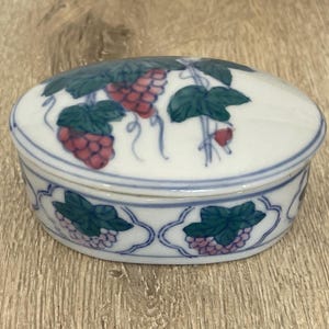 以下が含まれることがあります： ピンクと紫のブドウが描かれた、青と緑のフローラルデザインの白い楕円形のセラミック製の小物入れ。蓋には、緑の葉とピンクのブドウがついたつるが描かれています。
