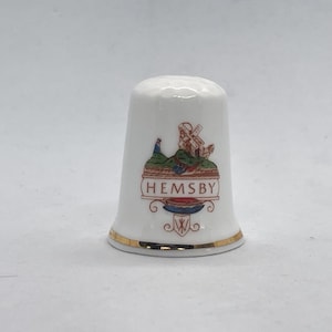 Peut inclure: Dé à coudre en céramique blanche avec un bord doré et un motif représentant un moulin à vent, une personne et le mot "HEMSBY" en rouge. Un objet de collection.