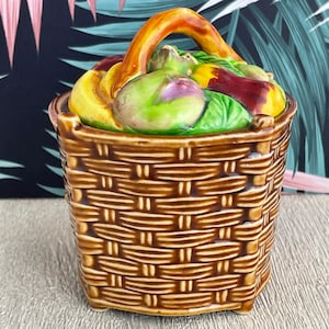 Peut inclure: Un récipient en céramique en forme de panier avec une texture tissée dans des tons bruns. Le panier est rempli de fruits et légumes colorés sculptés, dont des bananes, des pommes et des aubergines. Il a une poignée incurvée de couleur orange chaud.