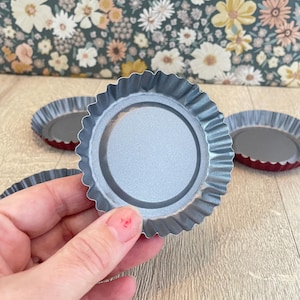 May include: Close-up of several small, silver-colored tart pans with crimped edges and a red base. The pans are round and have a fluted design around the rim. The background features a floral pattern.