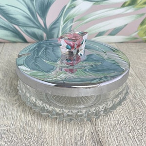 May include: A clear glass jar with a silver rim and lid, topped with a decorative square handle. The lid has a floral design. The jar sits on a wooden surface.