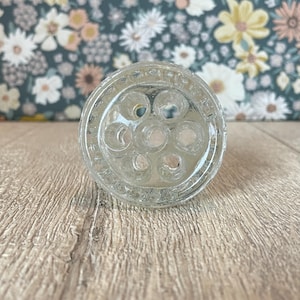 May include: Clear glass flower frog with multiple holes for arranging stems. The round frog has a textured rim and is designed to hold flowers in place. A vintage floral arranging tool.