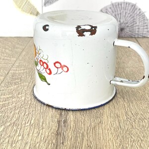 May include: White enamel mug with a blue rim and a floral design featuring red berries and yellow flowers.