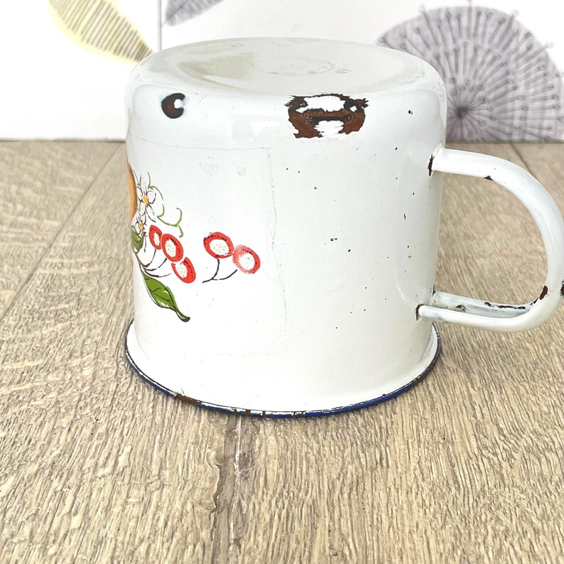 May include: White enamel mug with a blue rim and a floral design featuring red berries and yellow flowers.