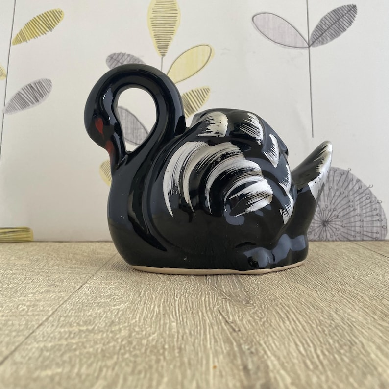 May include: A black ceramic swan figurine with white brushstrokes on its body and a red accent on its beak. The swan has a curved neck and is sitting on a light-colored surface.