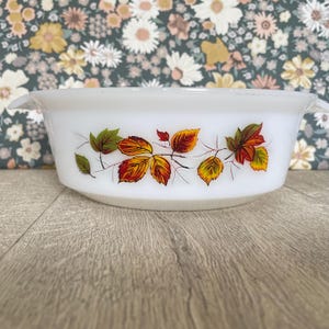 May include: White oval casserole dish with handles, decorated with a colourful autumn leaf design. The leaves are in shades of red, orange, yellow, and green. A vintage kitchenware item.