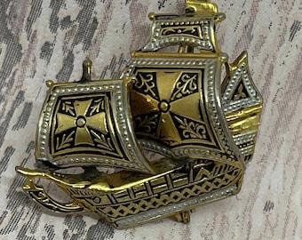 May include: A vintage ship brooch with gold and silver tones. The brooch features intricate detailing, including a cross design on the sails and a decorative hull. The ship's design is reminiscent of a historical vessel.