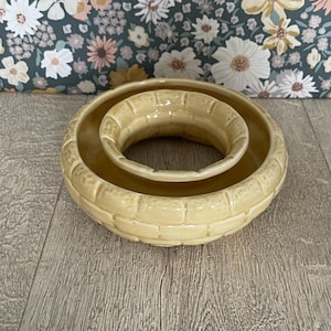 May include: A pale yellow ceramic bowl with a raised brick-like texture on the exterior and a central circular opening. The object features a glossy finish and a rounded, donut-shaped design, presenting a consistent light yellow colour throughout.
