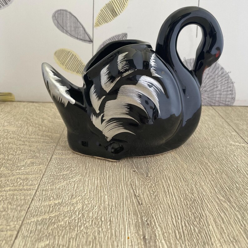 May include: A black ceramic swan-shaped planter with silver painted details. The swan has a long neck and a curved head, with a red dot for an eye. The planter has a wide opening for plants or other decorative items.