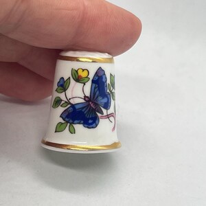 May include: A white porcelain thimble with a gold rim and a blue butterfly design. The butterfly is depicted with intricate details, and the thimble also features green leaves and a yellow flower. A decorative sewing accessory.